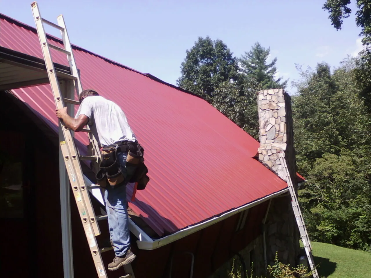 Roofer on a ladder beside a red metal roof and stone chimney — Soffit & Fascia Repair by Roofing Brothers in Deerfield